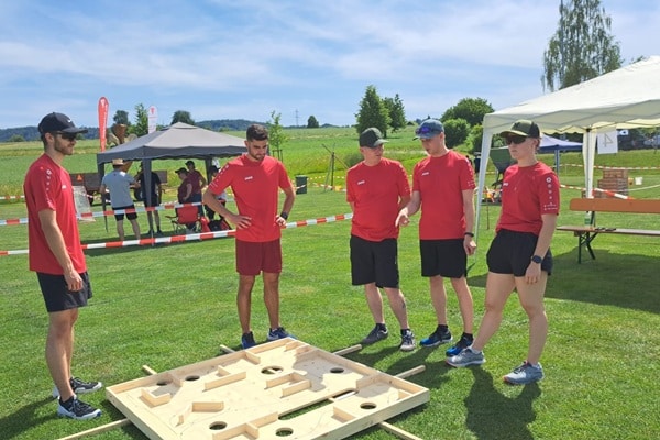 Fünf Personen in roten T-Shirts spielen ein großes Holz-Geschicklichkeitsspiel auf einer Wiese bei einem Outdoor-Event